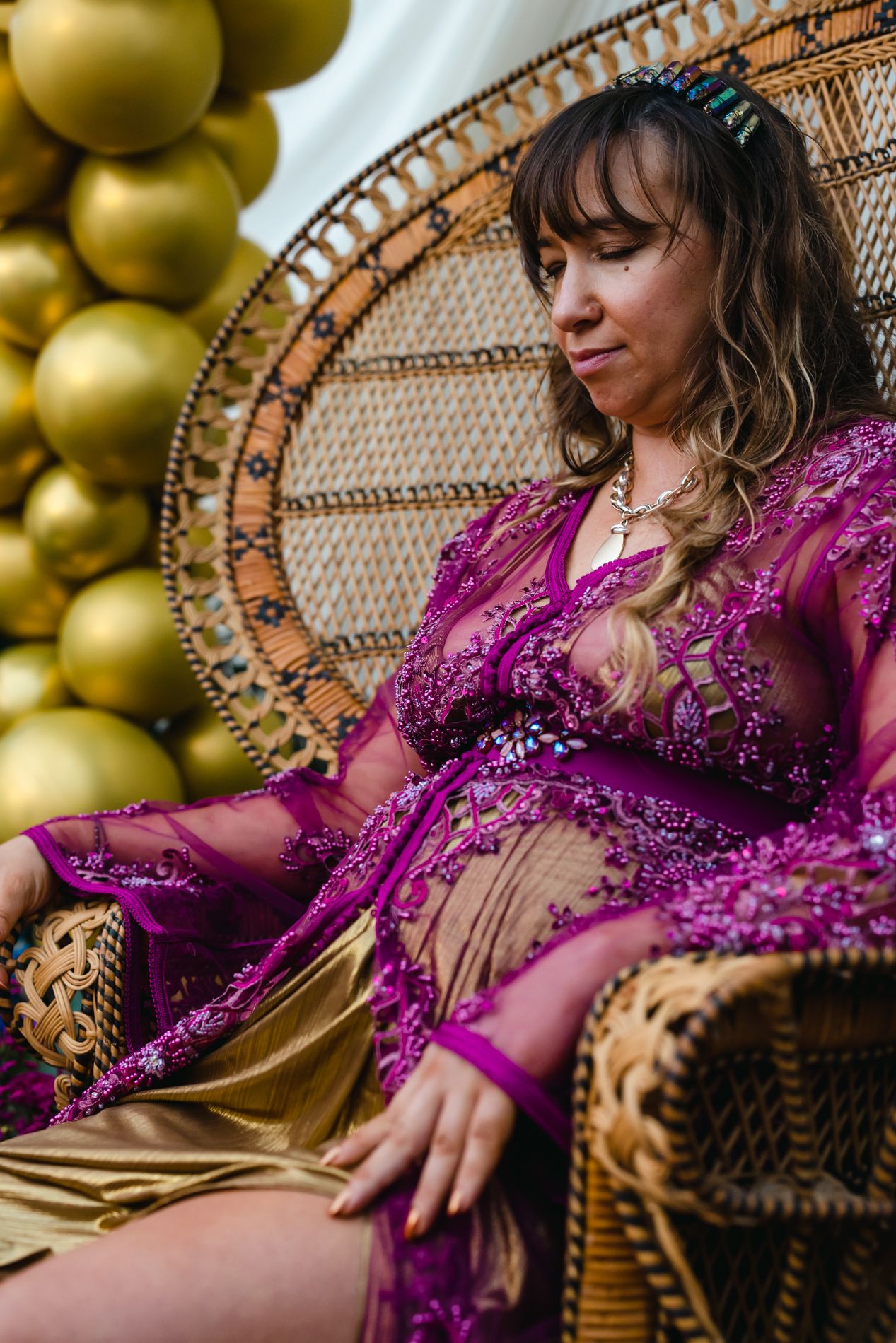 Mother-to-be in a beaded magenta robe seated on a wicker peacock chair with gold balloons — styled maternity shoot by Murrieta Media in Southern California